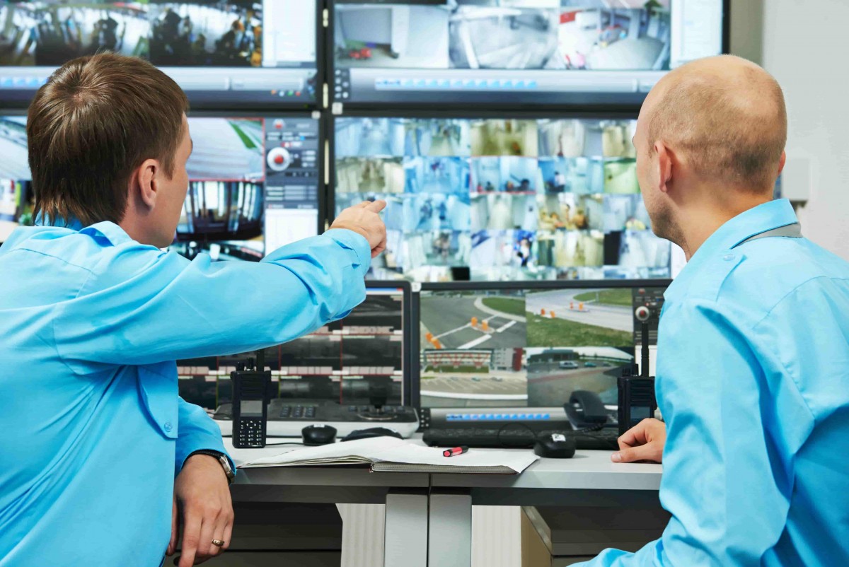 Photo de dos agents de sécurité vu de dos à un bureau muni de plusieurs ordinateurs devant un mur d'écrans de télésurveillance